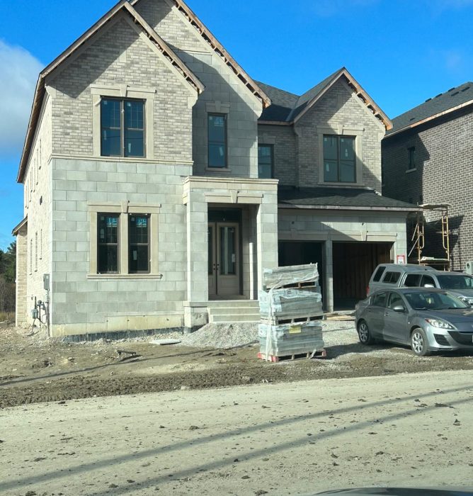 A large two-story house under construction with stone blocks on the lower level, brick on the upper level, and stacks of building materials in the dirt driveway.